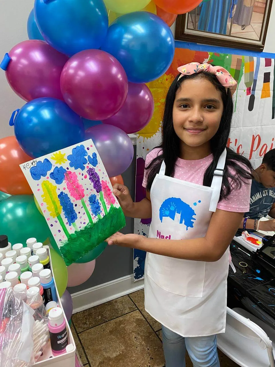 A young maker showing a bright flower painting and a Nashville Crafting Club apron.