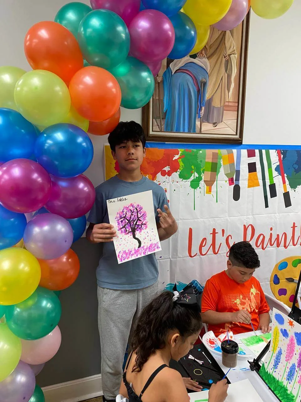 A teenage artist showing a purple tree painting beside a balloon garland.